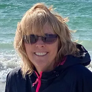 A woman wearing sunglasses and a jacket standing on the beach.