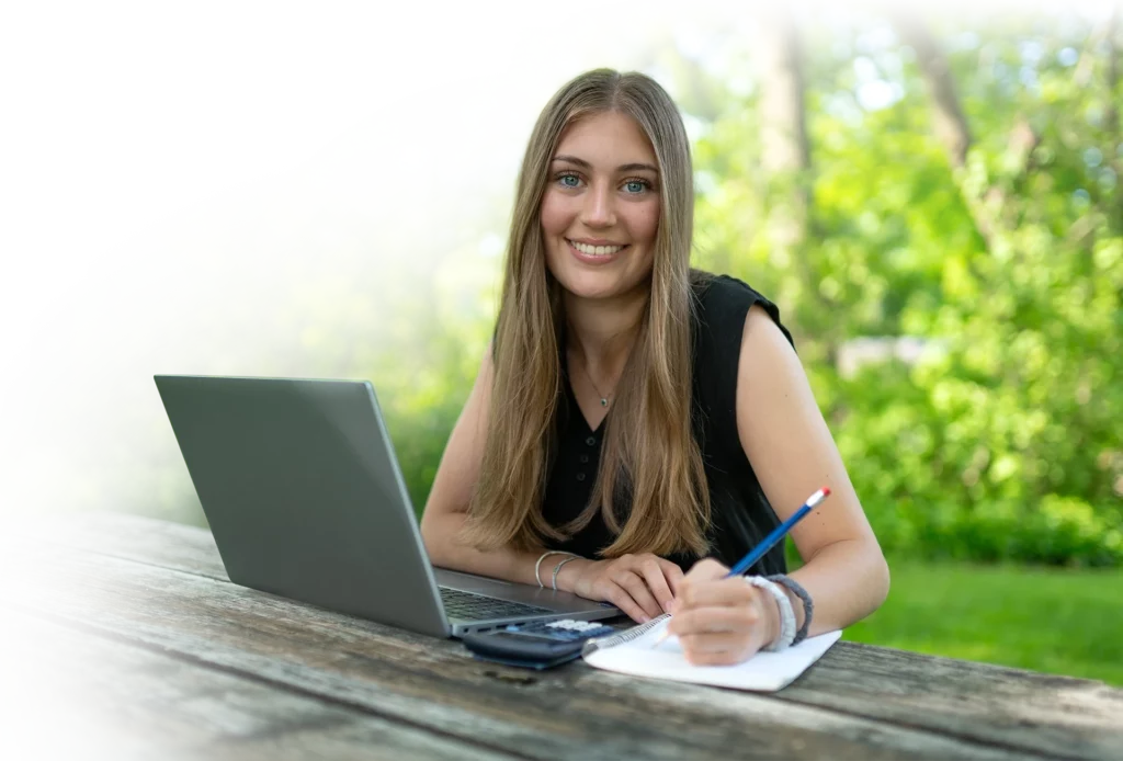 Smiling young lady sitting at a picnic table with a laptop, calculator, paper and holding a pencil.